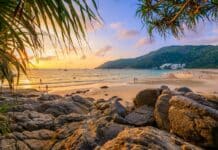 Atardecer en Nai Harn Beach, Phuket, con cielo naranja, nadadores, gente en la playa de arena, veleros en el mar y rocas a lo largo de la orilla.